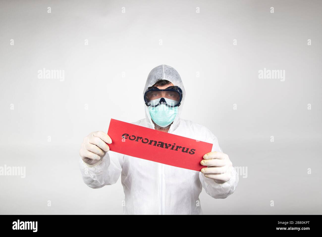 Uomo con maschera chirurgica, occhiali, tuta protettiva bianca e segno rosso con la parola coronavirus isolato in studio su sfondo bianco. Nuova pandemia epi Foto Stock