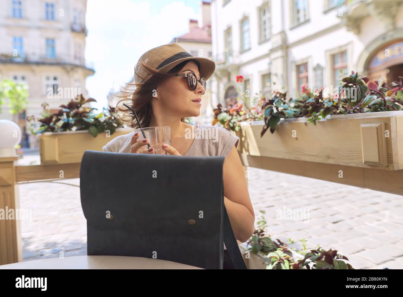 Bella donna di mezza età in occhiali da sole, cappello bere bevande fredde in estate caffè all'aperto, donna seduta da sola, sorridente guardando lontano Foto Stock