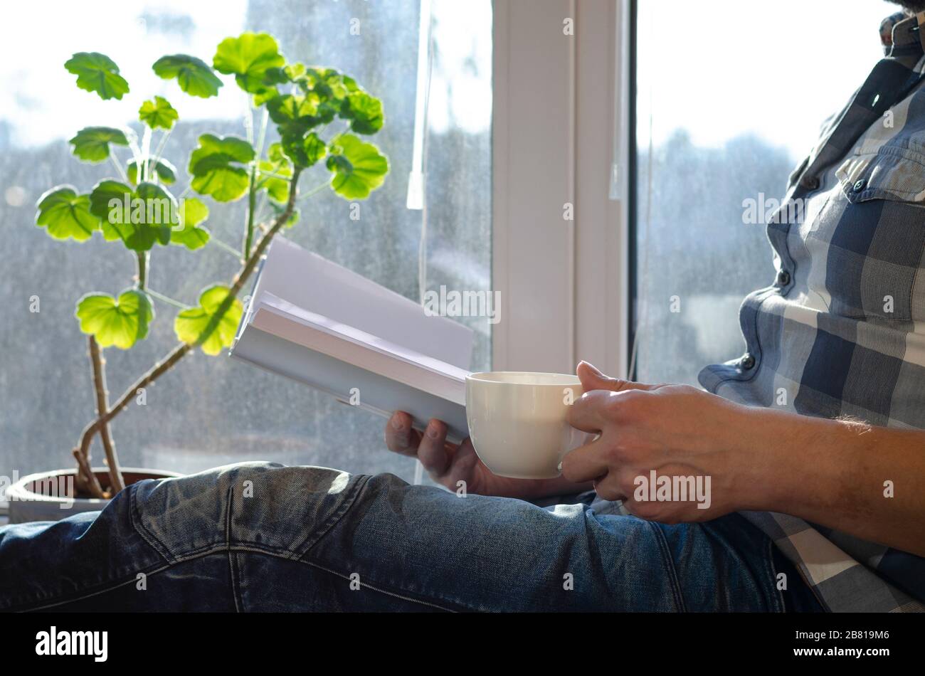 Un uomo siede su un davanzale con una tazza di caffè e un libro durante la quarantena Foto Stock