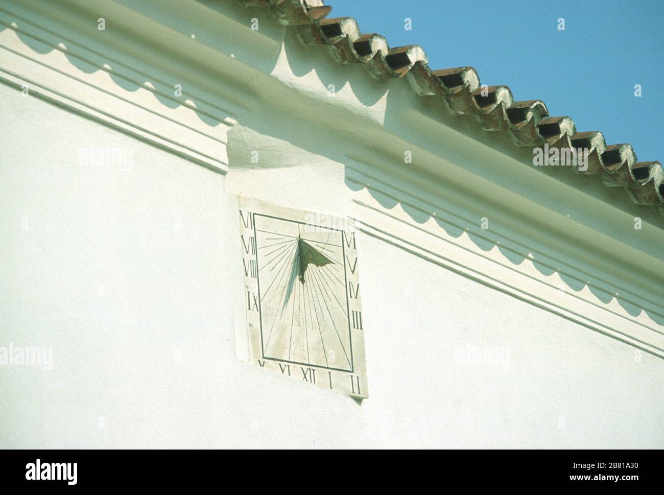 Meridiana con numeri romani, montata in alto sulla parete bianca in pietra di un edificio a Parga, Preveza, Epiro, Grecia. Il tetto piastrellato sopra getta un'ombra sulla parete. Foto Stock