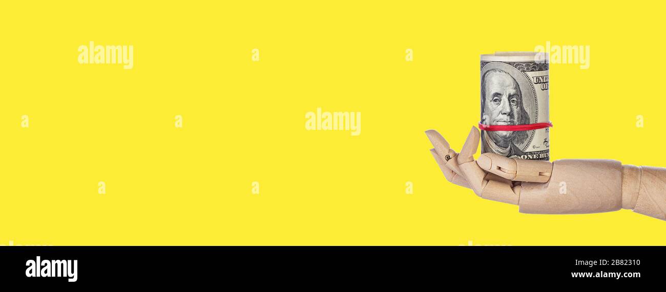 Mano di legno con denaro isolato su sfondo giallo. Rotolo di cento fatture del dollaro. Banconote da 100 USD, valuta in dollari americani. Ampio banner lungo Foto Stock