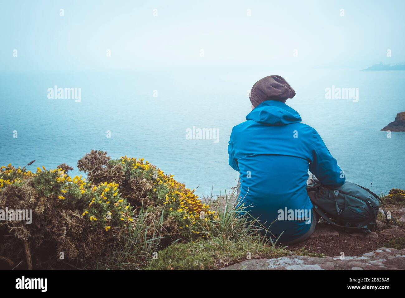 Giovane uomo con zaino, impermeabile e cappello lanoso seduto sul bordo di una scogliera costiera guardando il mare in una giornata piovosa e maledetta. Immagine con toni esterni w Foto Stock
