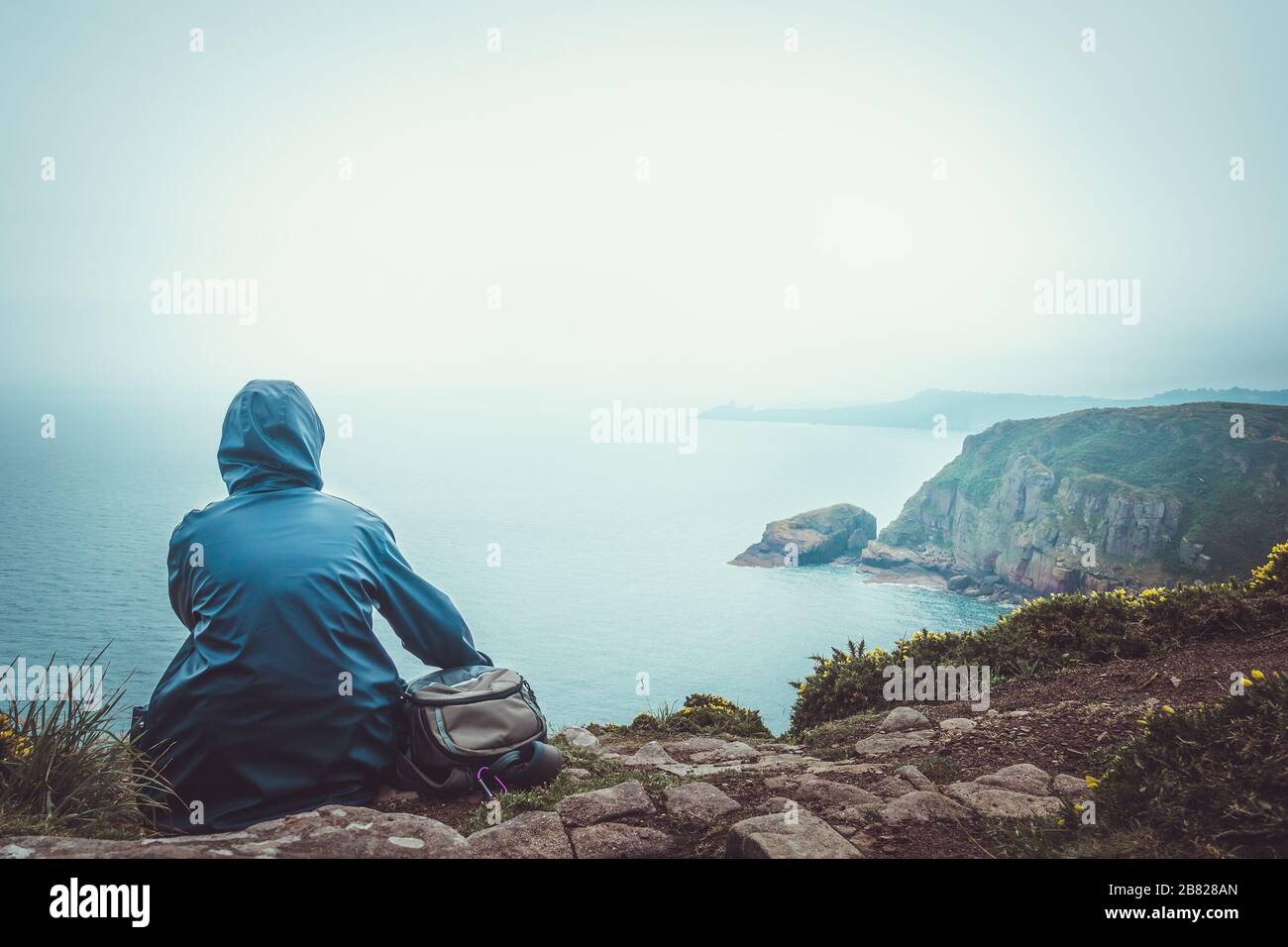 Donna con impermeabile e zaino seduto sul bordo di una scogliera che guarda il mare in una giornata piovosa e maledetta. Immagine concettuale a toni all'aperto con copie Foto Stock