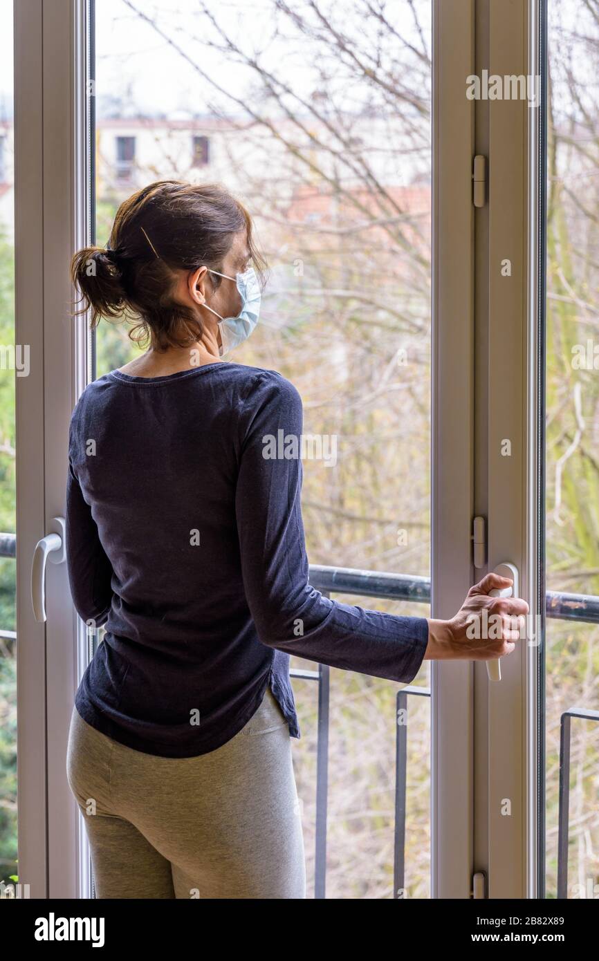 Una donna in quarantena a casa, che indossa una maschera medica per il viso e un abbigliamento casual, è in piedi inattivo di fronte a una finestra chiusa, fissandosi nello spazio. Foto Stock