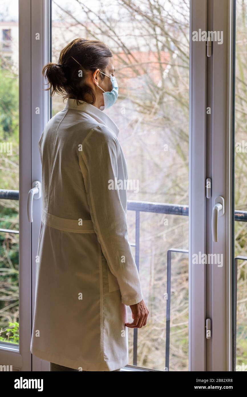 Un infermiere che indossa una maschera medica per il viso e un cappotto bianco, è in piedi inattivo davanti a una finestra chiusa, fissandosi nello spazio. Foto Stock