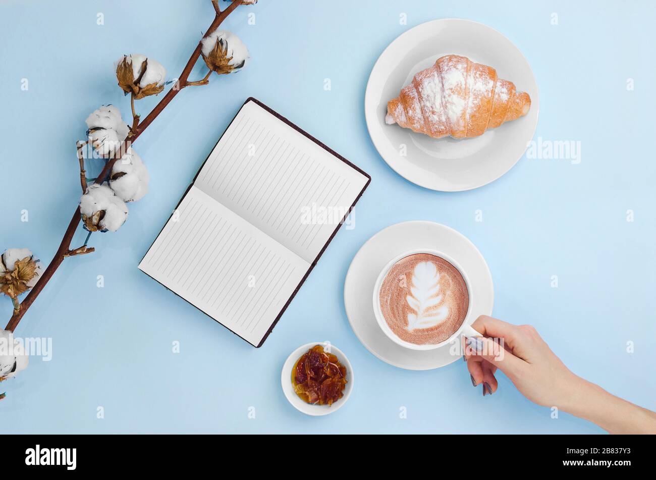 Mani che tengono caffè con latte art e taccuino con croissant, marmellata e branca di cotone Foto Stock