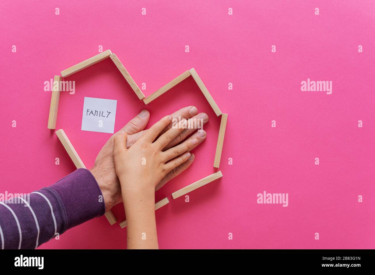 Mano di un padre e di un bambino accatastati uno sopra l'altro all'interno di una forma a cuore fatta di pioli di legno. Su sfondo rosa con il segno Famiglia. Foto Stock