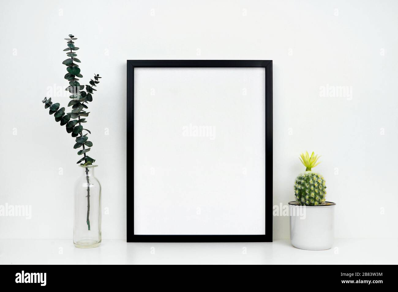Mokup telaio nero con cactus e rami su uno scaffale o scrivania. Ripiano e parete bianchi. Orientamento verticale della cornice. Foto Stock