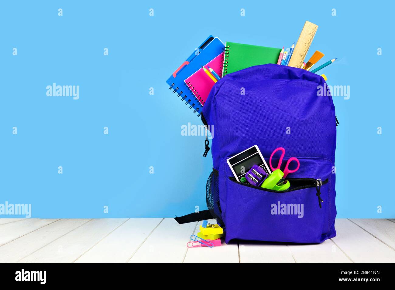 Zaino viola pieno di forniture scolastiche su sfondo blu. Torna al concetto di scuola. Spazio di copia. Foto Stock