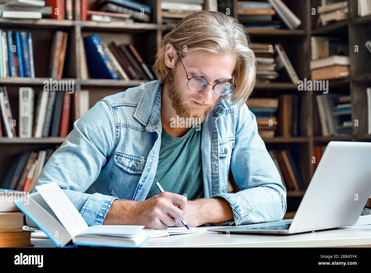 Giovane studente concentrato maschio alla biblioteca scrivania moderno notebook scrivere in notebook. Foto Stock