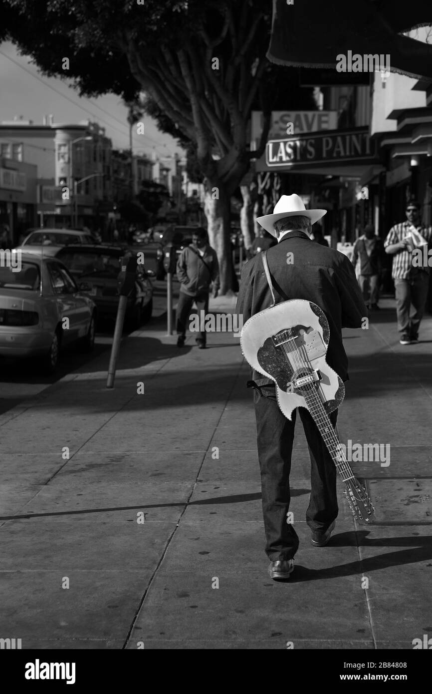 Un uomo in un cappello cowboy testi mentre porta una chitarra slung sopra la sua schiena camminando lungo Mission Street, San Francisco, California, Stati Uniti Foto Stock