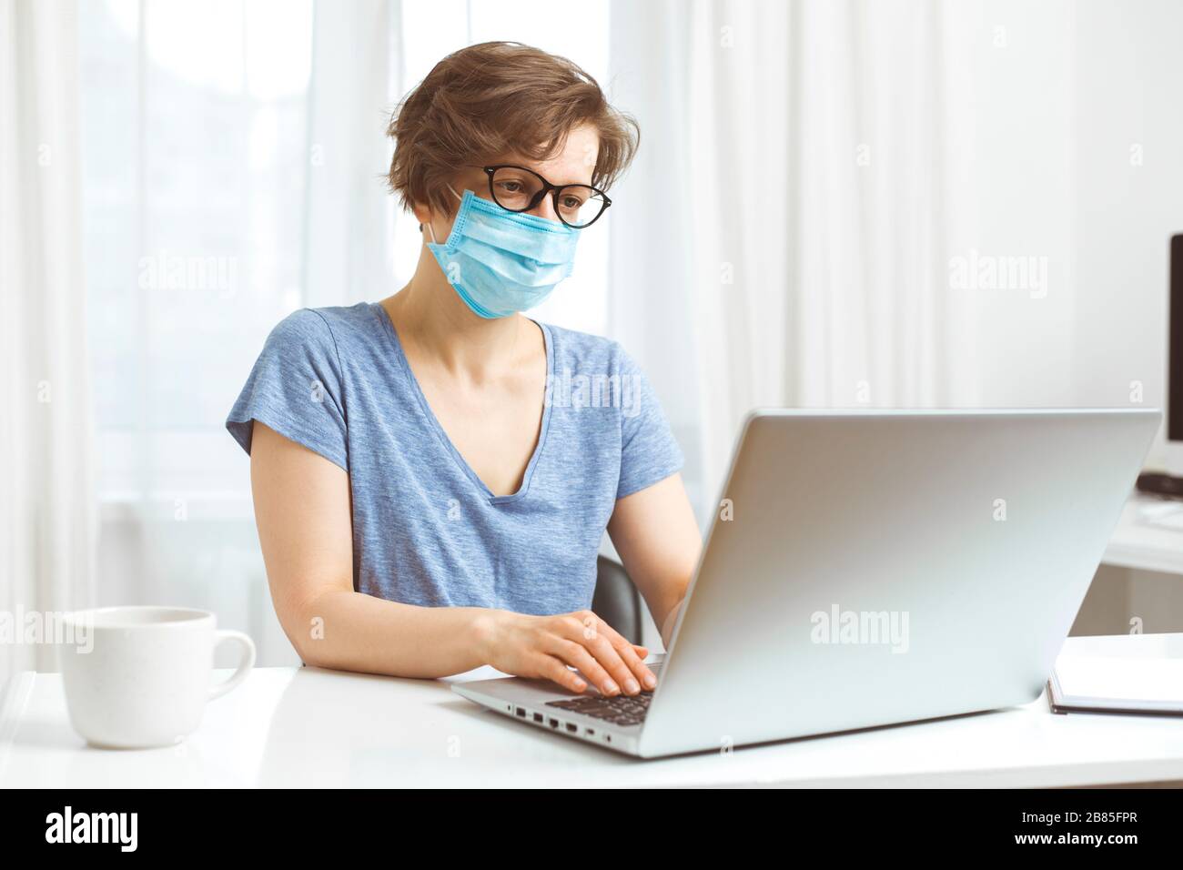 Una donna freelancer in una maschera medica lavora da remoto a casa su un computer. Il concetto di quarantena durante le malattie virali. Foto Stock