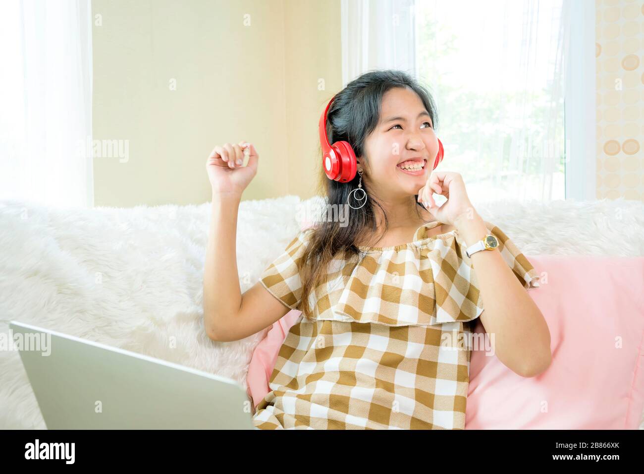 Le donne asiatiche hanno un aspetto carino indossando cuffie rosse e ascoltando musica collegata al notebook. Nella sua camera da letto. Seduta sul divano con una luce bianca Foto Stock