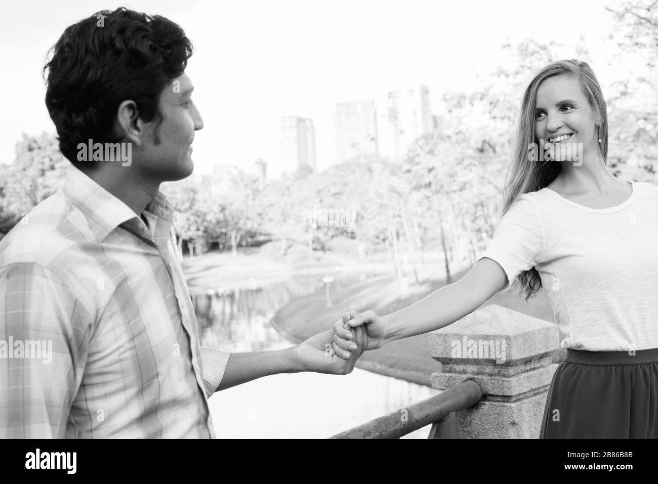 Felice coppia multietnica sorridente mentre si guarda l'un l'altro e tenendo le mani insieme sul ponte del parco Foto Stock