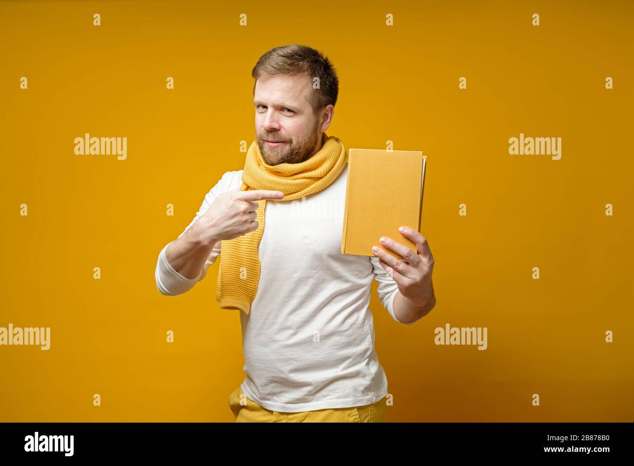 Uomo carino in una sciarpa, tiene un libro giallo in mano, punta ad esso con il dito indice e guarda la macchina fotografica. Foto Stock