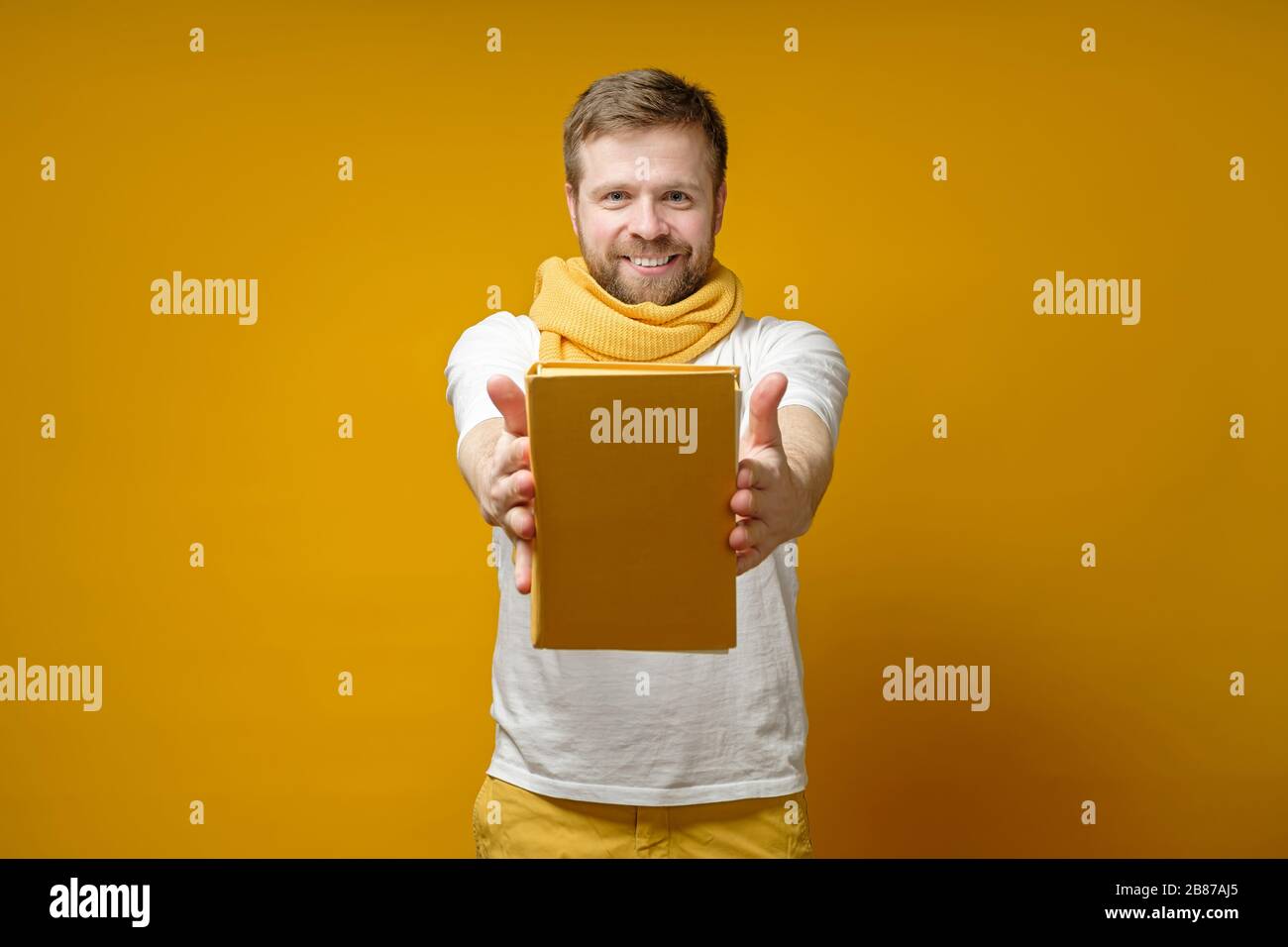 L'uomo soddisfatto in una sciarpa gialla tiene un libro sulle mani allungate, lo offre e sorride alla macchina fotografica. Concetto di istruzione. Foto Stock