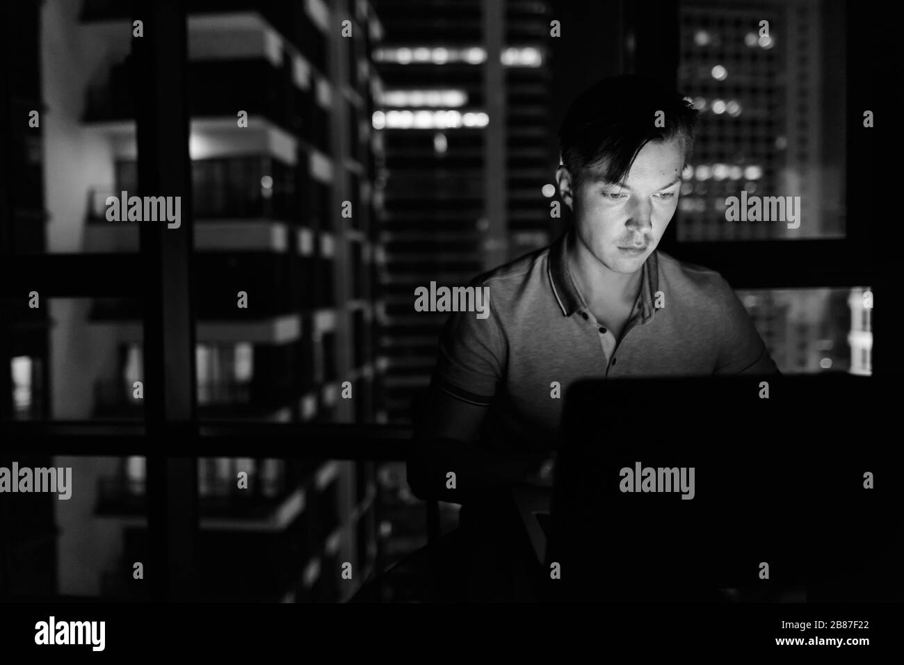 Giovane bell'uomo che usa il laptop contro la finestra di vetro con vista della città di notte Foto Stock