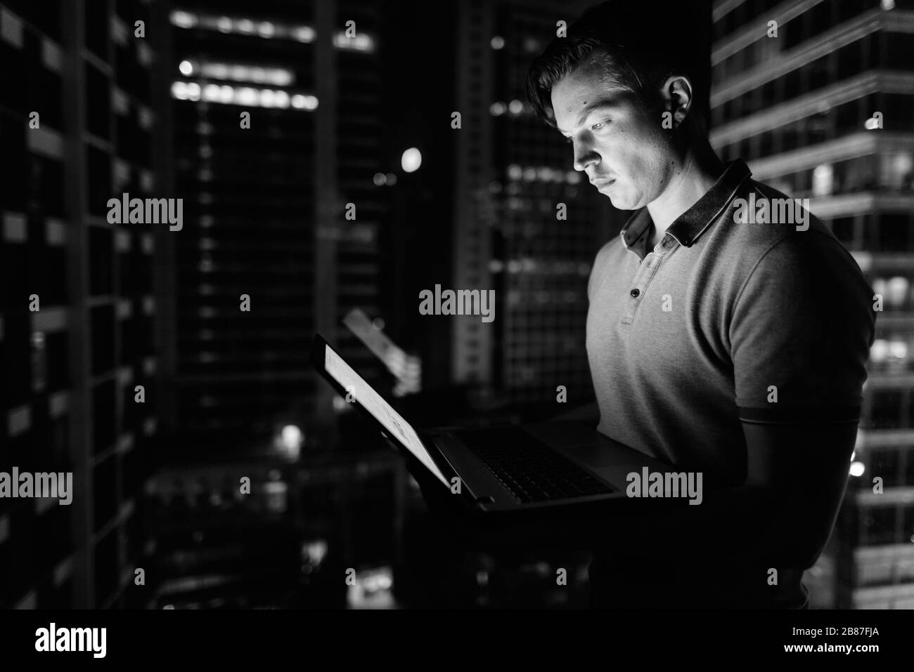 Giovane bell'uomo che usa il laptop contro la finestra di vetro con vista della città di notte Foto Stock