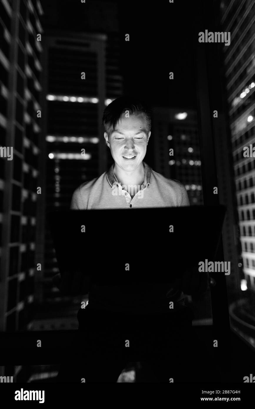 Giovane bell'uomo che usa il laptop contro la finestra di vetro con vista della città di notte Foto Stock