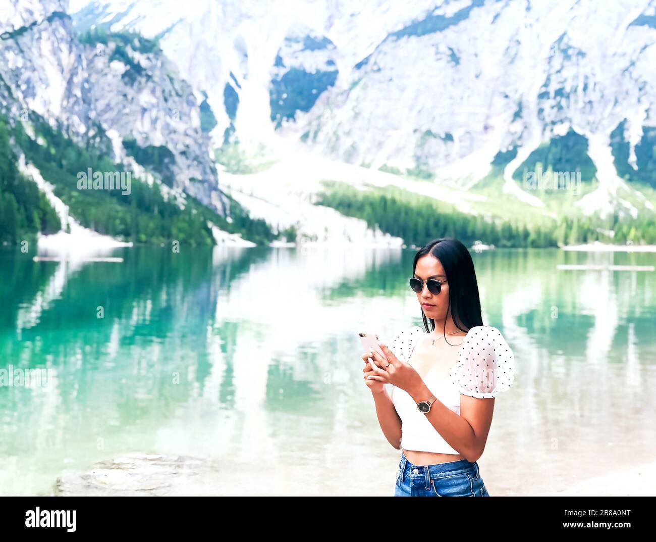 Bella giovane viaggiatore che usa smartphone sul lago nelle Dolomiti mentre viaggia in estate con incredibile vista del lago Braies sul retro Foto Stock