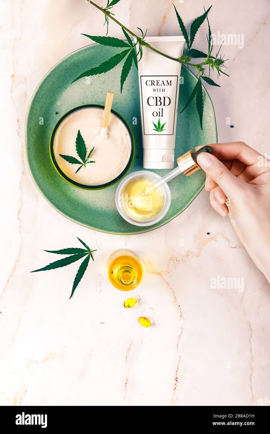 Pipettare con olio cosmetico CBD in mani femminili su fondo tavolo con crema, foglie di cannabis Foto Stock Pipettare con olio cosmetico CBD in mani femminili su fondo tavolo con crema, foglie di cannabis Foto Stock