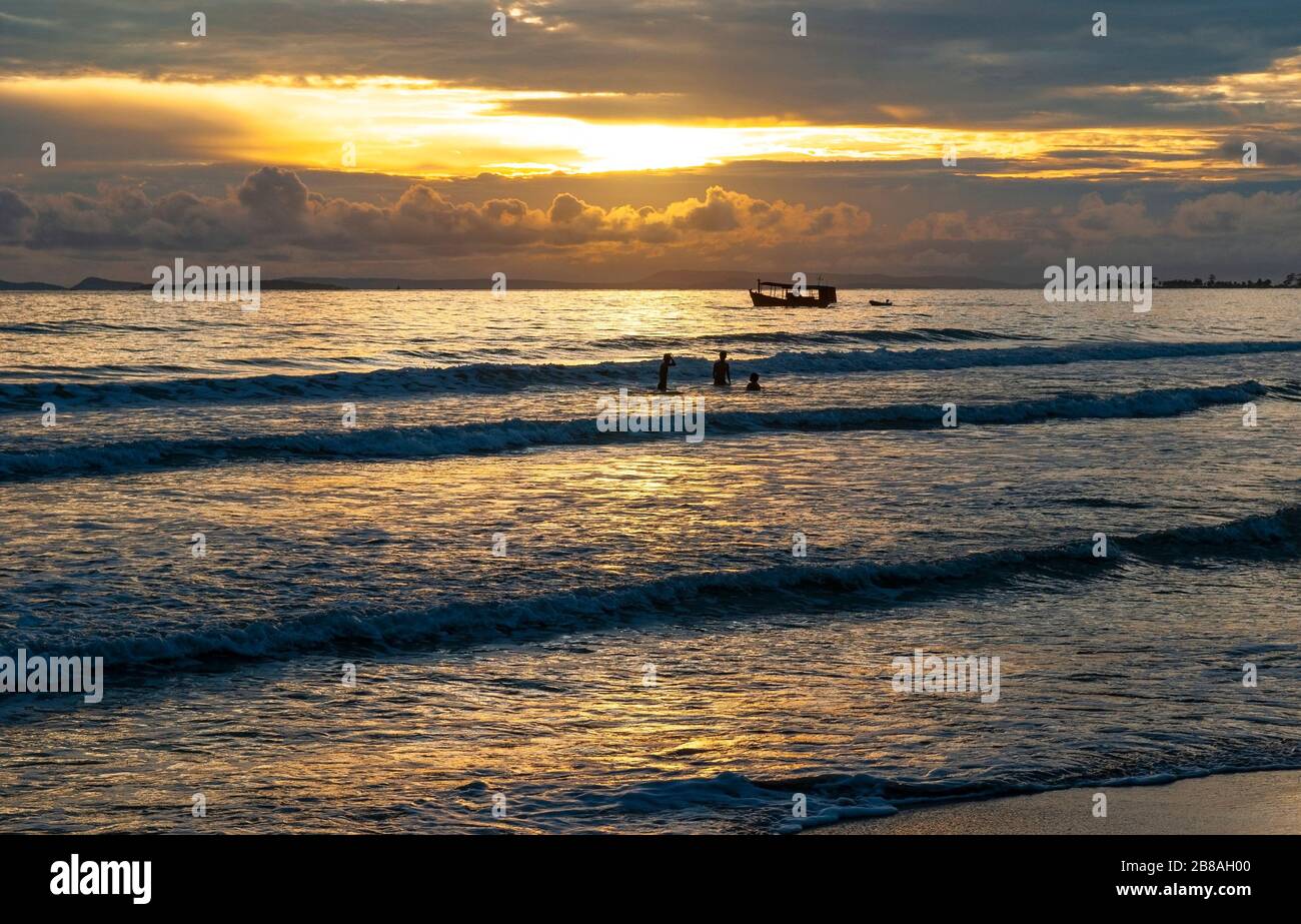 Tramonto lungo la spiaggia di Otres a Sihanoukville con la silhouette di persone che giocano nelle onde e l'ombra di una barca da pesca, Golfo della Thailandia, Cambogia. Foto Stock
