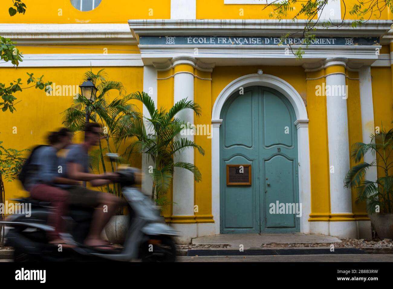 Pondicherry, India - 17 marzo 2018: Coppia irriconoscibile su uno scooter che passa davanti ad un edificio giallo della scuola con sfocatura del movimento Foto Stock