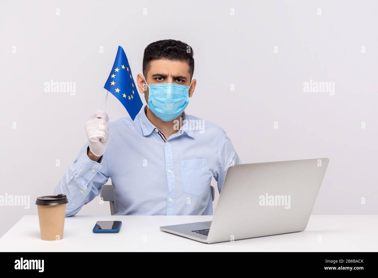 Ufficiale in maschera igienica e guanti in possesso della bandiera UE, preoccupata, avvertimento di epidemia di coronavirus in Europa, quarantena e misure preventive f Foto Stock