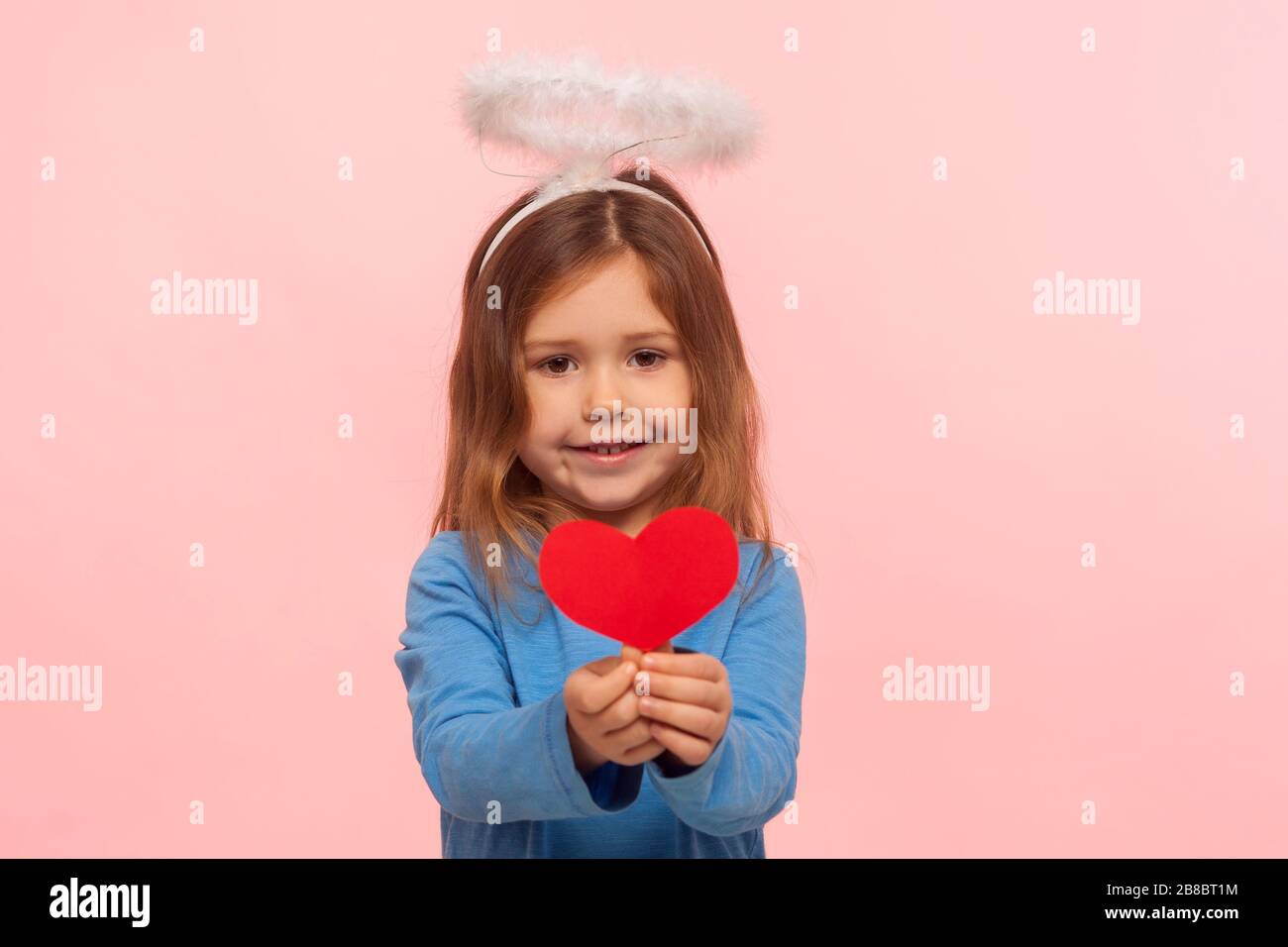 Ritratto di adorabile ragazza prescolare carina con angelic nimbus tenendo carta cuore e sorridente a macchina fotografica, piccolo cupido per San Valentino condividere l'amore. Foto Stock