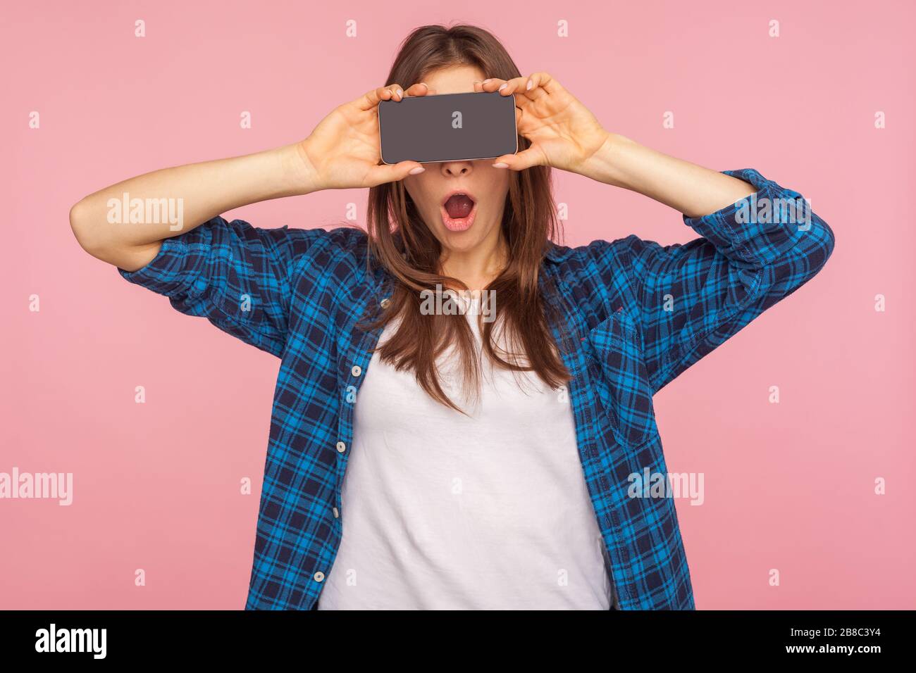 Pubblicità incredibile su dispositivi mobili! Ragazza stupita in camicia a scacchi che copre gli occhi con smartphone e tenendo la bocca aperta a sorpresa, nascondendo h Foto Stock