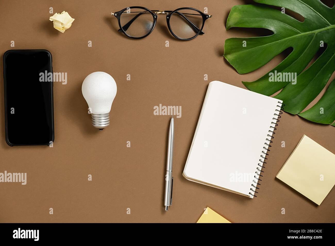 Idee ispirazione concetti con lampadina, penna, smartphone moderno, foglie di mostera, occhiali, notebook con spazio copia su sfondo marrone. Modello Foto Stock