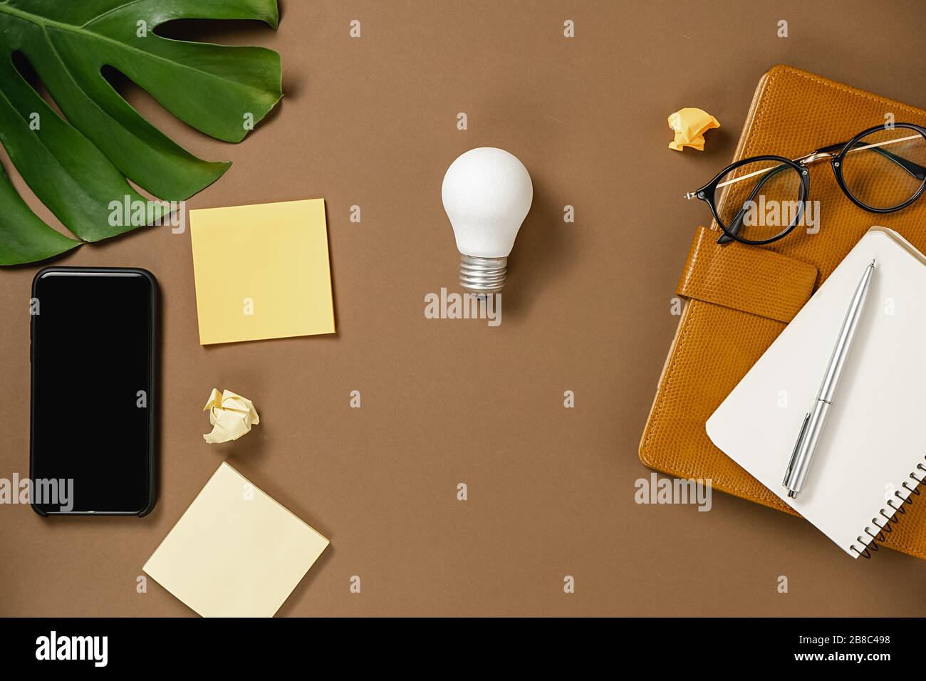 Creative vista dall'alto piano di lavoro con penna, foglie di mostera, occhiali, notebook, moderna composizione smartphone con spazio copia su sfondo marrone Foto Stock