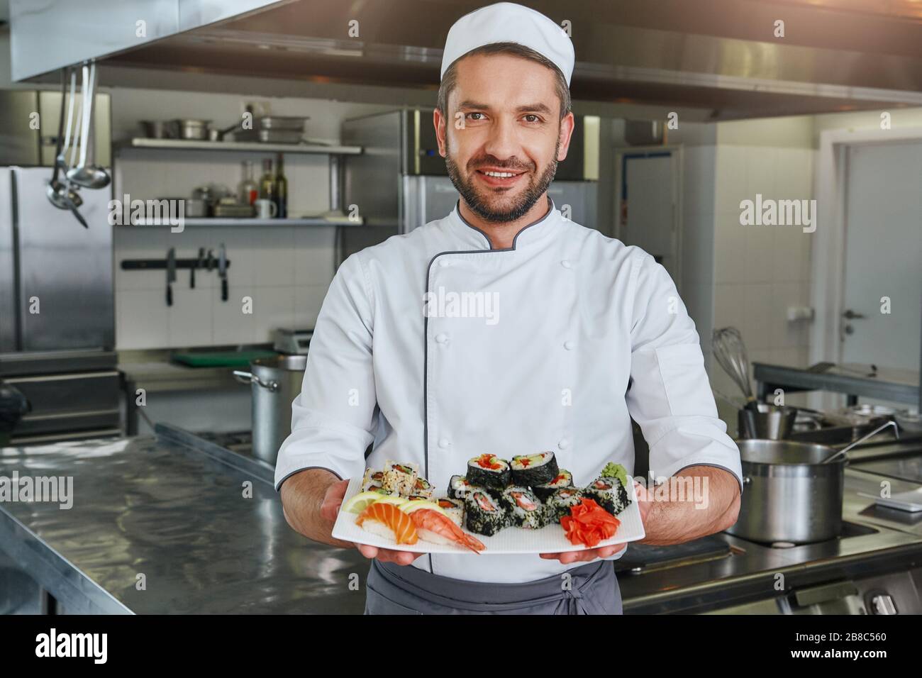 Il maestro del sushi ha preparato il sushi al ristorante. Sta in piedi in cucina commerciale e tenendo il suo piatto mentre guarda la macchina fotografica con un sorriso. Vista frontale Foto Stock