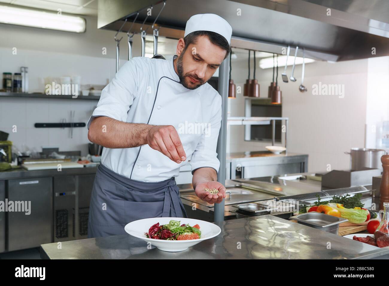 Cuoco caucasico dai capelli scuri che indossa l'insalata di cottura in uniforme bianca. Stagiona l'insalata per renderlo migliore Foto Stock Cuoco caucasico dai capelli scuri che indossa l'insalata di cottura in uniforme bianca. Stagiona l'insalata per renderlo migliore Foto Stock