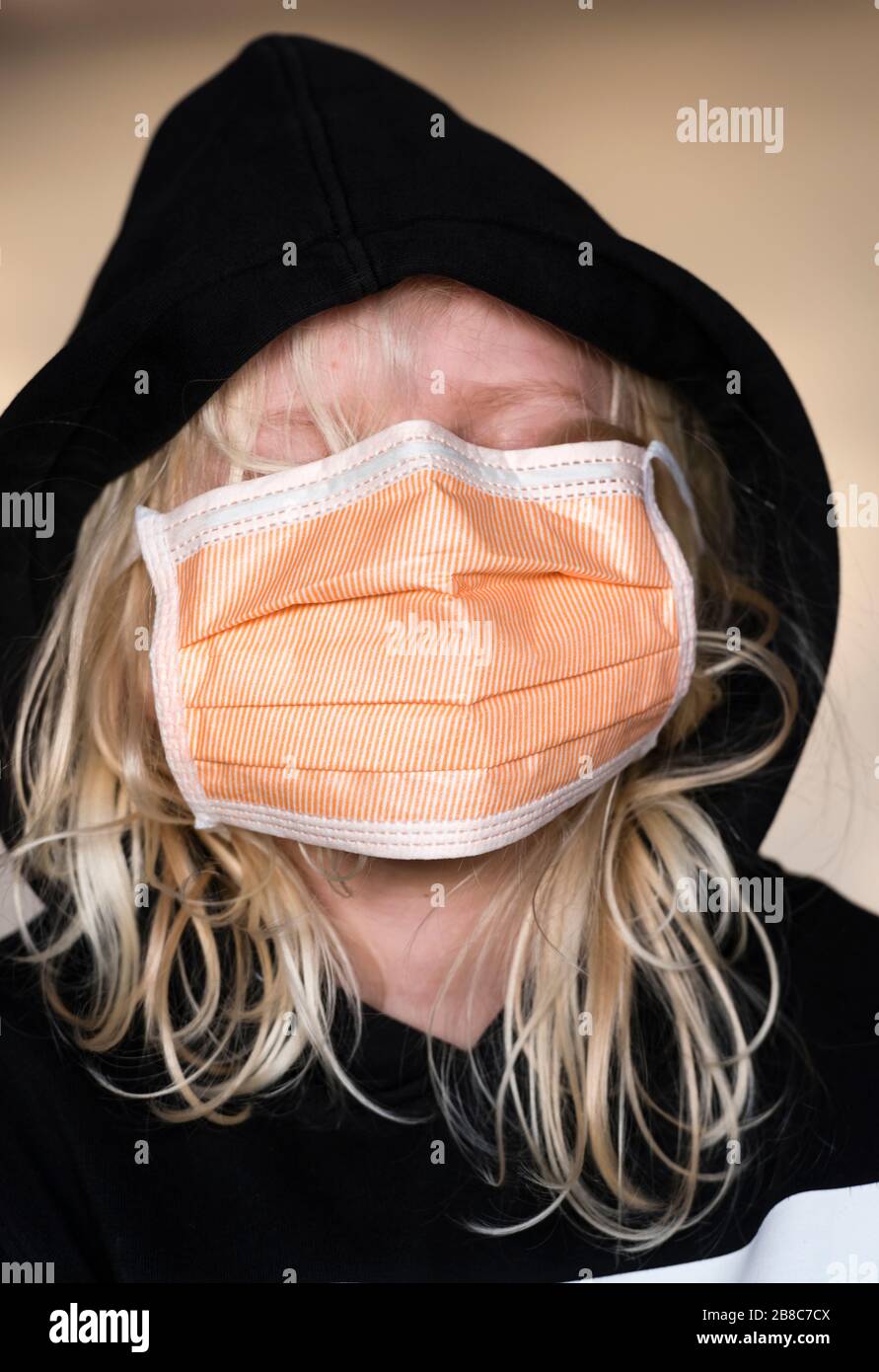 ragazzo di 12 anni con lunghi capelli biondi indossa una maschera facciale di protezione dalle infezioni e una felpa con cappuccio nera. Etnia caucasica, faccia coperta completamente da fa Foto Stock