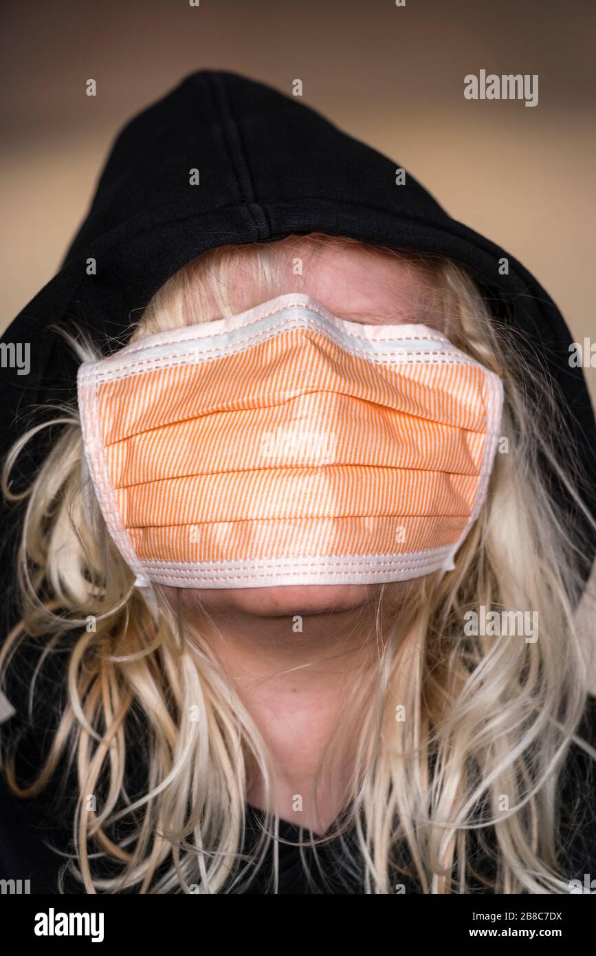 ragazzo di 12 anni con lunghi capelli biondi indossa una maschera facciale di protezione dalle infezioni e una felpa con cappuccio nera. Etnia caucasica, faccia coperta completamente da fa Foto Stock