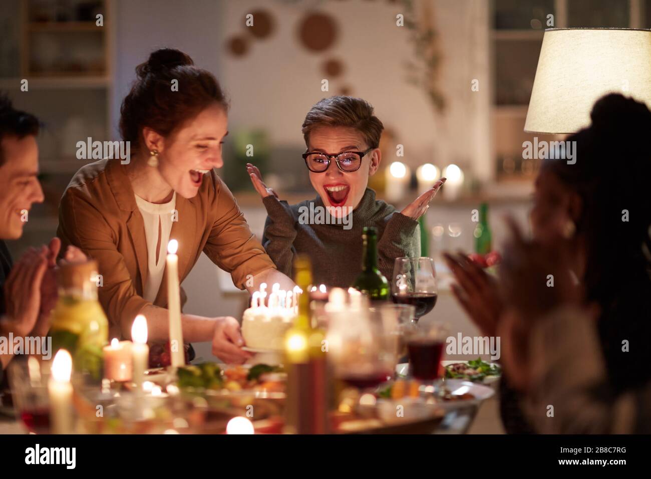 Felice giovane donna in occhiali è così eccitato di torta di compleanno con candele mentre si siede al tavolo insieme con i suoi amici Foto Stock