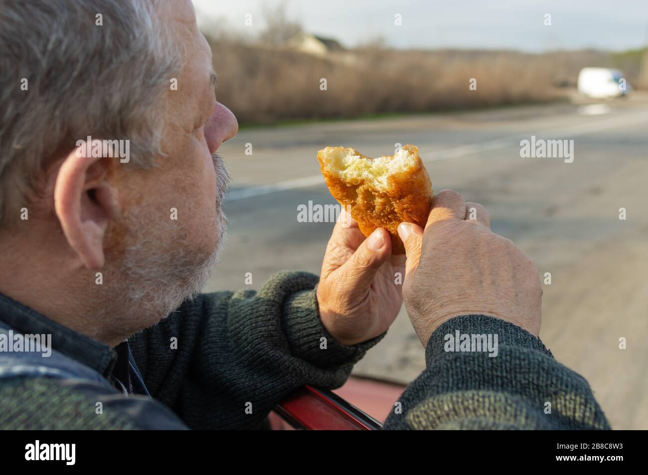 Ritratto del driver superiore affamato di Caucasian che mangia la pattuglia appoggiando i suoi gomiti sulla porta dell'automobile Foto Stock