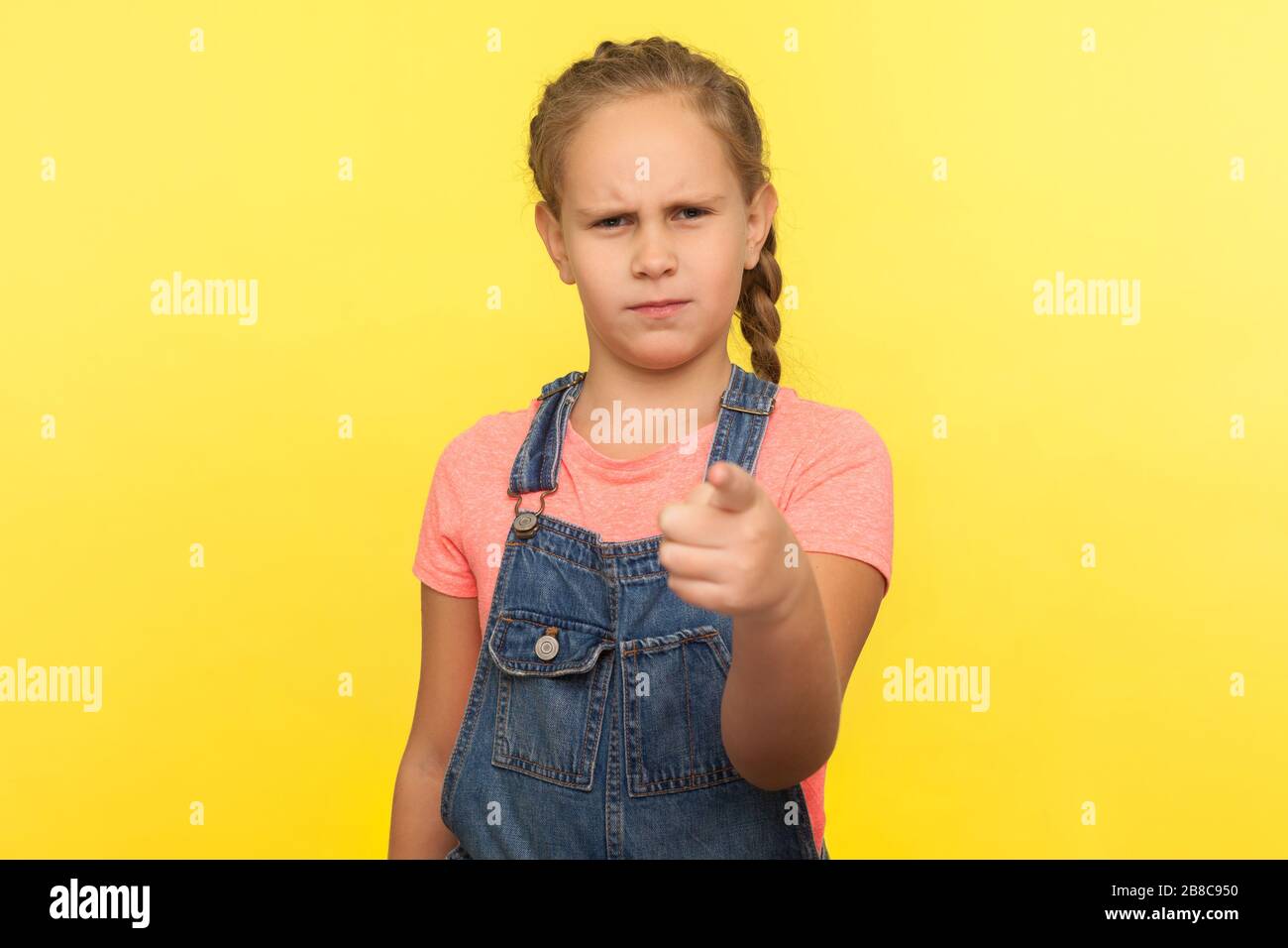 Ehi! Ritratto di bambina bossy con treccia in tuta denim che punta alla macchina fotografica e che guarda con espressione dispiaciuta arrabbiata, facendo la scelta. In Foto Stock