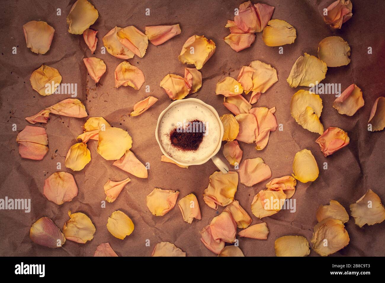 Tazza di caffè con crema e cannella a forma di cuore con petali rosa su carta kraft sbriciolata. Disposizione piatta. Composizione centrale. Vignettatura. Foto Stock