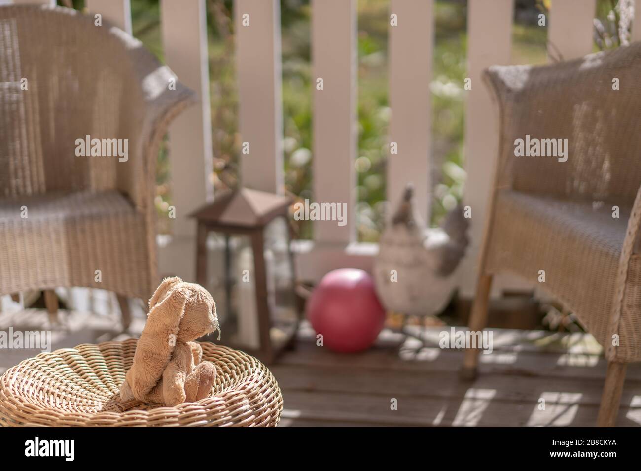Giocattolo ripieno coniglio pasquale seduta su una sedia di vimini sulla veranda al sole, guardando la palla rosa. Vista ad alta angolazione, primo piano, concetto. Foto Stock