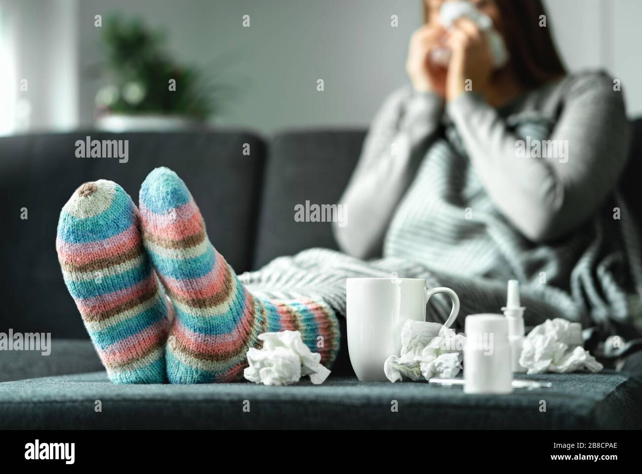 Donna malata con influenza, freddo, febbre e tosse seduta sul divano a casa. Persona malata che soffia il naso e starnutisce con tessuto e fazzoletto. Calzini in lana. Foto Stock