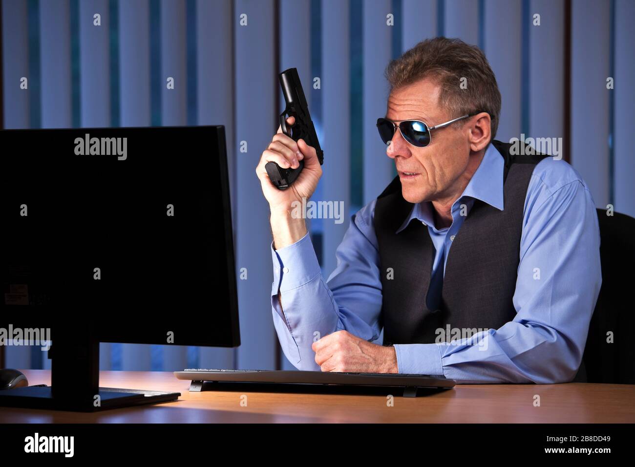 Uomo d'affari criminale seduto con la pistola in mano ad una scrivania con monitor in un ufficio buio di notte Foto Stock