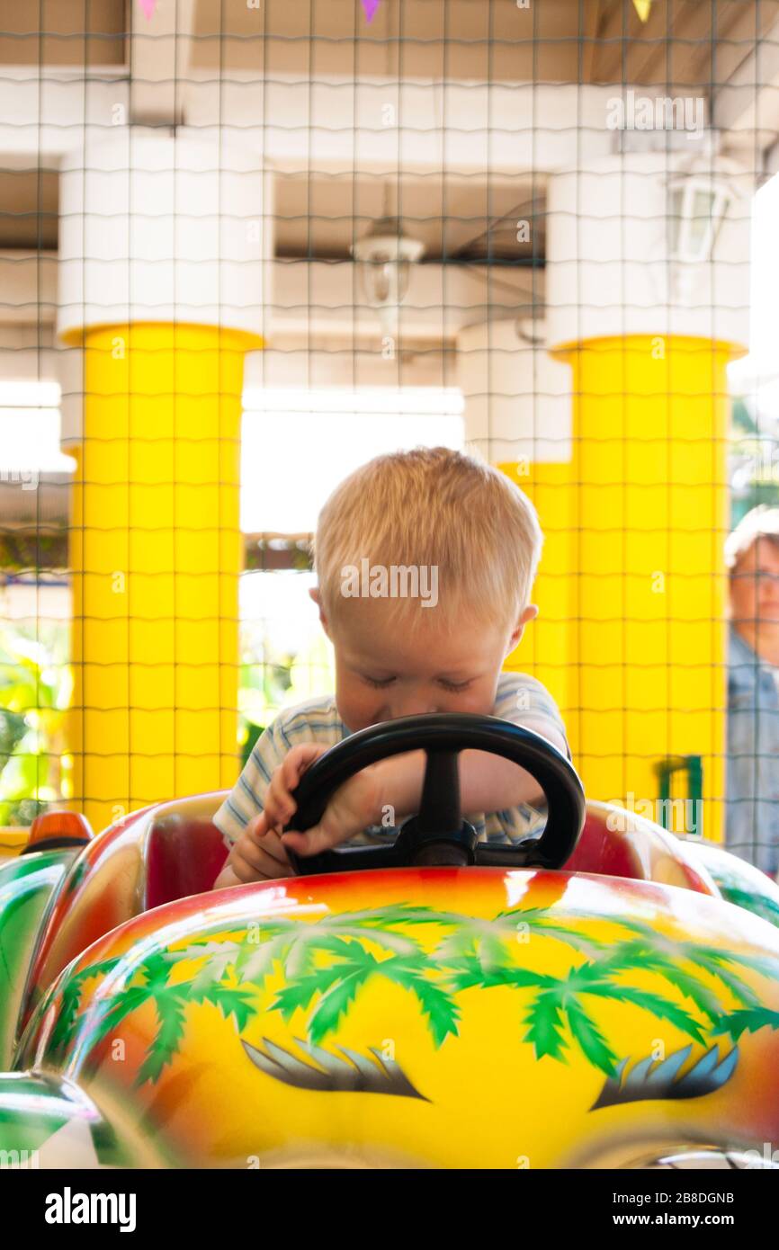 Un ragazzino biondo molto gioioso e felice di tre anni è in sella a un'auto gialla con un'auto verde nel parco divertimenti estivo Riviera, Sochi con la schiena allo spettatore. Riposo, tropici, riposo, attivazione, sorriso. Foto verticale Foto Stock