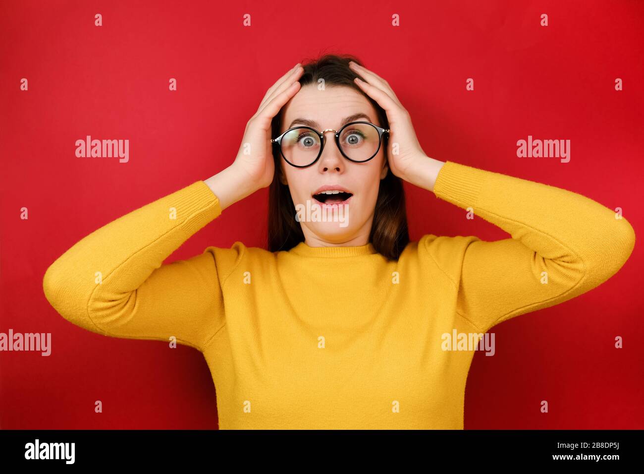 La donna spaventata Emotion guarda con espressione omg, tiene le mani vicino alla testa, indossa occhiali trasparenti, maglione giallo, isolato su sfondo rosso studio. Foto Stock