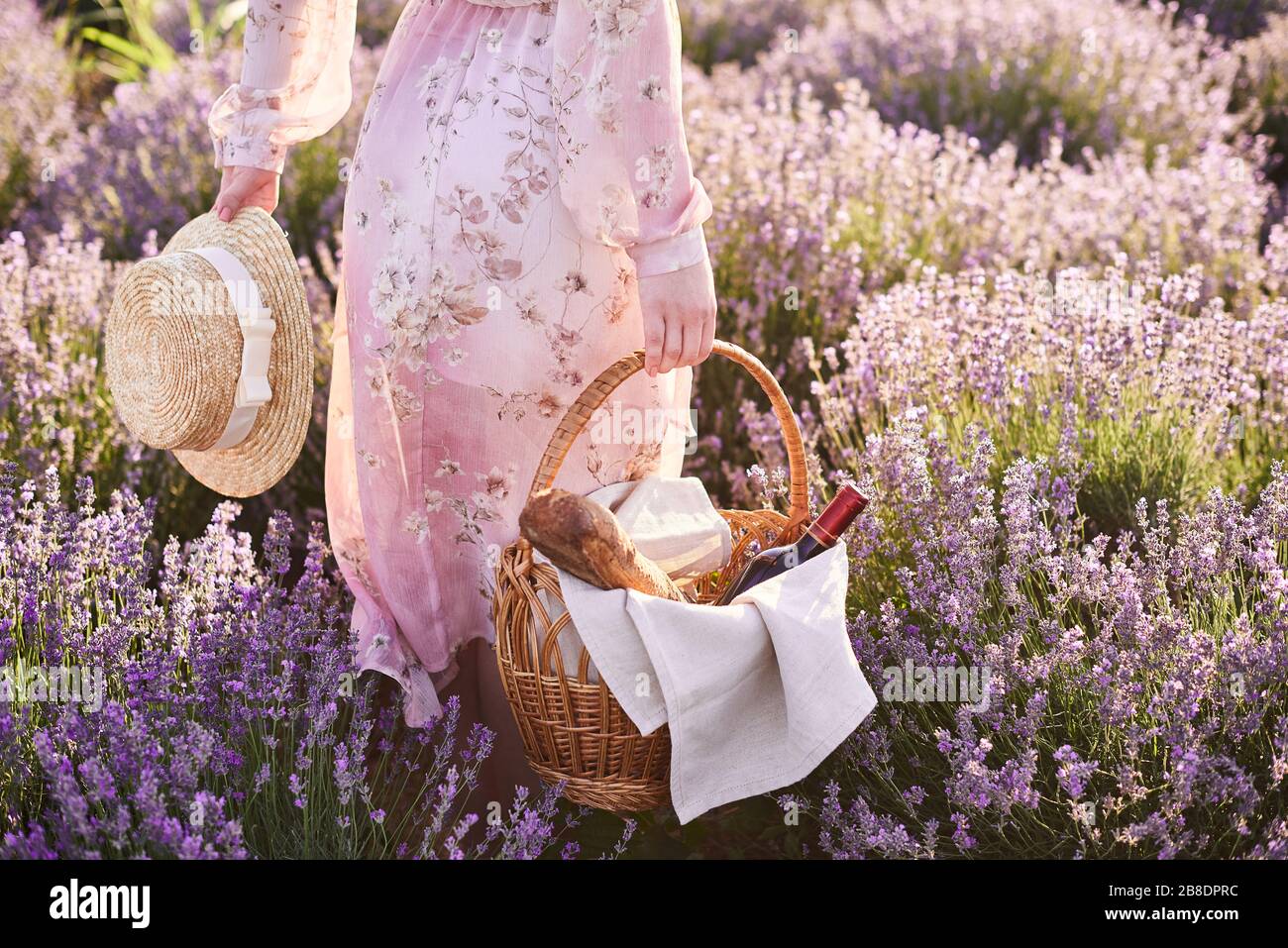 ragazza nel vestito romantico con un cappello di paglia in mano cerca un buon posto per picnic in campi di lavanda Foto Stock