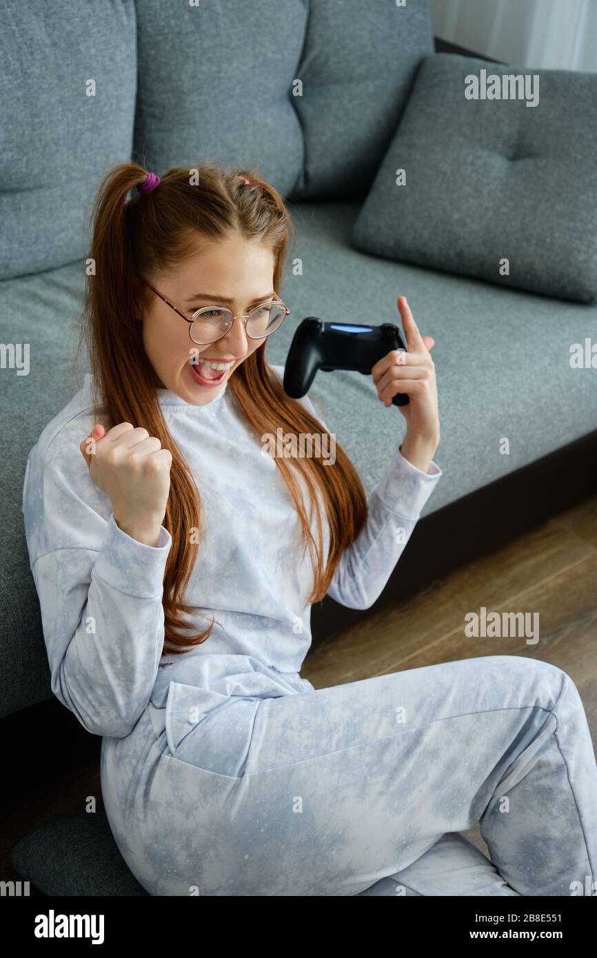 Una ragazza dai capelli rossi con occhiali, con lunghe code, si siede sul pavimento con gioia alzando le mani al petto con una console di gioco Foto Stock