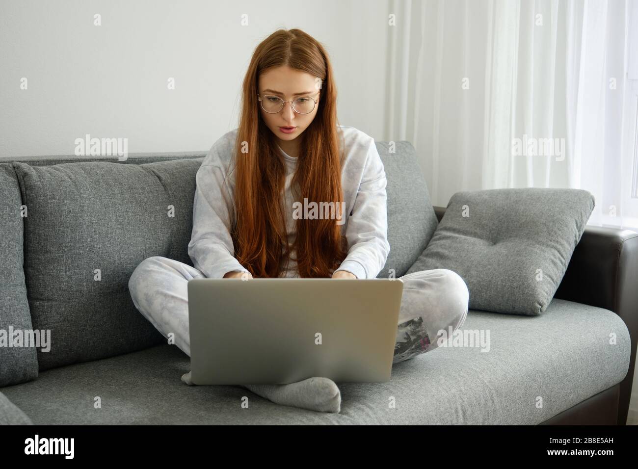 Una ragazza dai capelli rossi con occhiali, con capelli lunghi in pigiama si siede a gambe incrociate sul divano e guarda intensamente il laptop Foto Stock