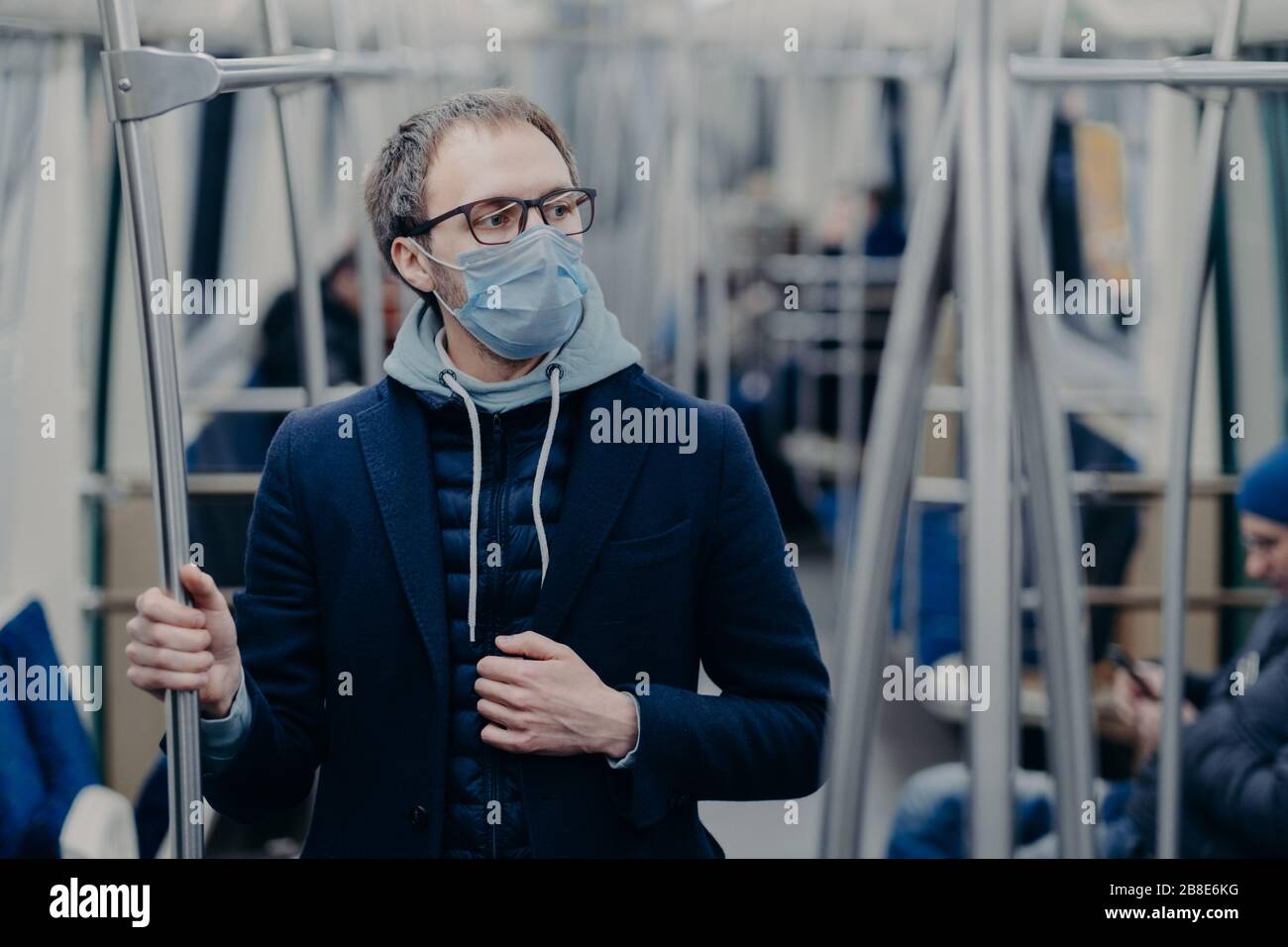 Il giovane uomo pensoso negli occhiali indossa la maschera chirurgica protettiva durante lo scoppio del coronavirus, pone nei mezzi di trasporto pubblici, pensa come superare le malattie Foto Stock