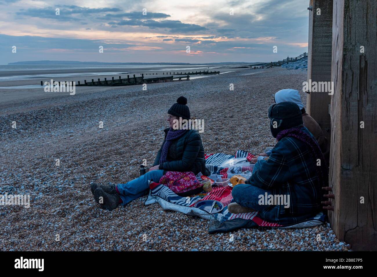 Una famiglia sedersi su una spiaggia fredda al tramonto, mangiare un pic-nic, per uscire durante le distanze sociali Foto Stock